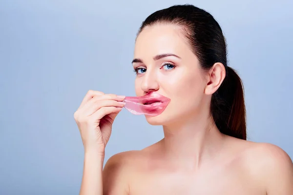 Young beautiful woman with natural make-up removing pink hydrogel patch from her lips. Korean cosmetics. Cosmetology and skin care. Isolated on grey. Beauty skin and anti-wrinkles care.