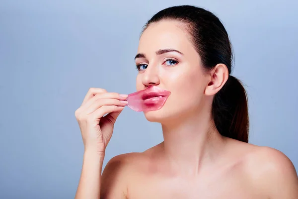 Young beautiful woman with natural make-up removing pink hydrogel patch from her lips. Korean cosmetics. Cosmetology and skin care. Isolated on grey. Beauty skin and anti-wrinkles care.