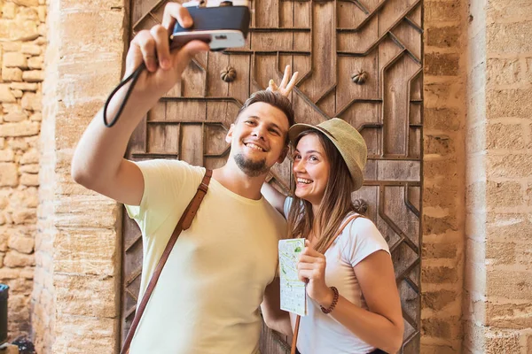 Portrait of happy and funny young couple of travellers taking selfie with film camera on vacations. Woman holding map in hands. Old wooden door on background.