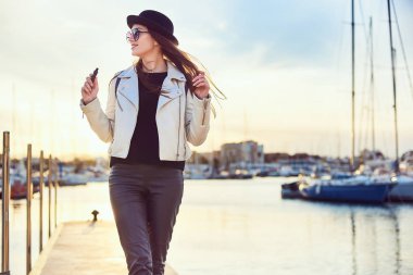 Genç ve güzel uzun saçlı kadın blogger vape, gülümseyen ve arka planda bir bağlantı noktası deniz ve deniz araçları ile gün batımında yürüyüş tutarak güneş gözlüğü yuvarlak siyah şapkalı.
