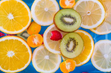 Taze ahududu, mandalina, kivi, portakal, limon ve portakal dilimleri mavi suda yüzer. Sulu ve renkli meyveler.