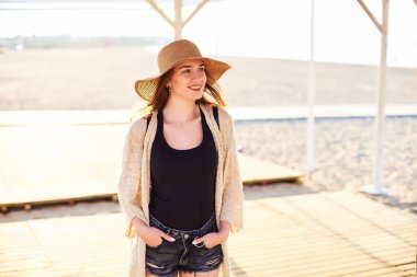 Elleri ceplerinde giymiş şık Bayan kot şort, tunik ve gülümseyen ve onun tatil yaz aylarında sahilde zevk hasır şapka sökük. Uzay kopyalayın. Seyahat ve Turizm kavramı.