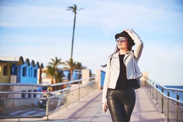 快乐的年轻迷人的女孩博客戴着黑色的帽子和圆形太阳镜正在海边散步 手里拿着手机 丹尼亚 西班牙 — 图库照片