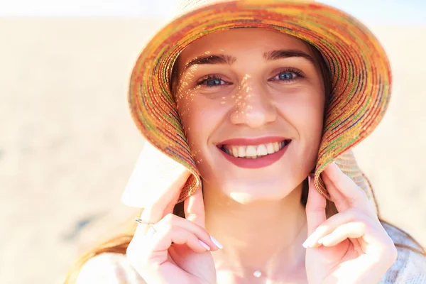 Portrait of smiling woman on beach wearing straw hat. Close up face of beautiful young woman feeling good at seaside. Happy girl wearing sun hat and looking at camera.