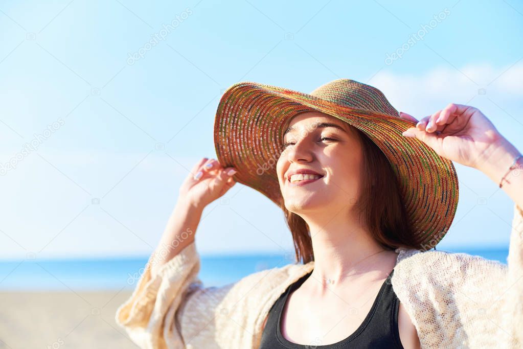 Portrait of young happy woman with closed eyes wearing straw hat relaxing and enjoying the sun on the beach. Close-up face of beautiful young woman feeling good at seaside.