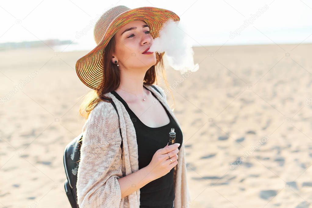 Young attractive happy woman wearing straw hat vaping electronic cigarette and blowing smoke on sunny summer day on the beach. Travel and tourism concept.