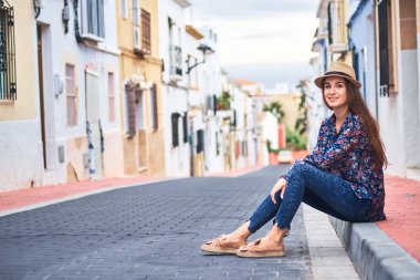 Denia İspanyol kasabasında küçük renkli binalar ile kaldırımda oturan genç gülümseyen kadın.