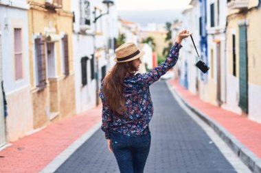 Arkadan görünüşü el eski fotoğraf makinesi ile sokakta yürürken şapkalı uzun saçlı bir kadın.