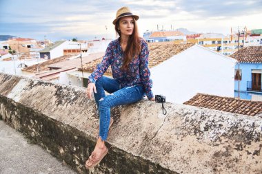 Genç gülümseyen uzun saçlı kadın turist onun tatil İspanya'da arka plan üzerinde renkli binalar ile küçük İspanyol şehrin panoramik manzaralı zevk şapkalı.