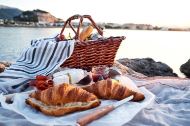 Güzel romantik piknik sepeti, kruvasan, kırmızı şarap, ekmek, reçel, peynir ve jamon sahilde denize yakın ile kompozisyon.