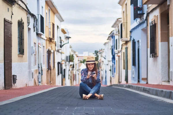 stock image Young smiling woman sitting on road and taking photos with old camera in small Spanish town.