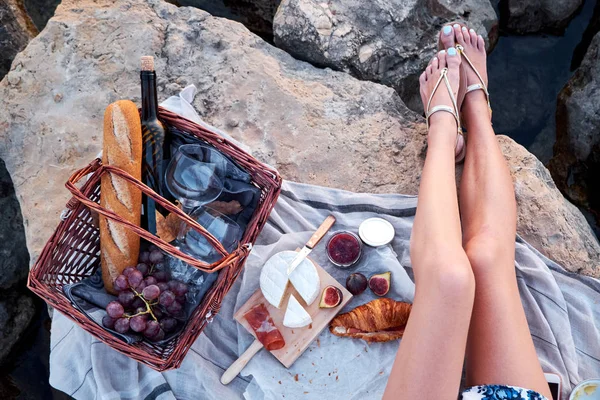 Top view of beautiful young woman sitting on the rocks with picnic basket with croissants, bread, jam, wine, glasses, cheese, figs and jamon.
