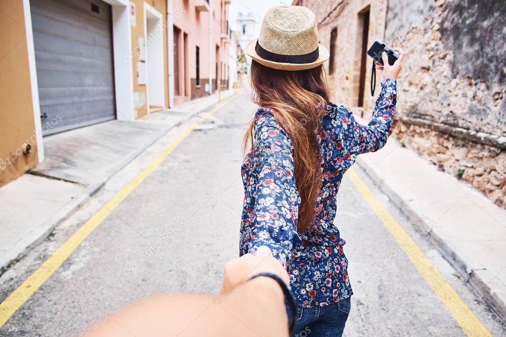 Back view of a beautiful young woman in hat holding man's hand and leading him on old town. Travelling and tourism concept.