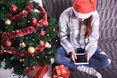 Hediye ile kanepede oturan ve tablet Noel ağacı yakın tutarak kazak ve kırmızı Santa şapka genç mutlu kadın. Yeni yıl ve Noel tatil kavramı.