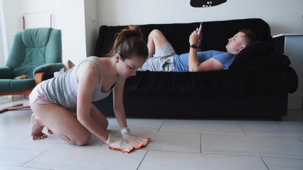 Zmęczony żona czyści podłodze płytki, a jej mąż leży na kanapie — Wideo stockowe