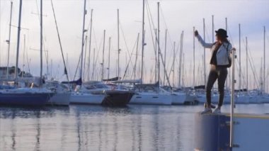 Mutlu genç çekici kız blogger siyah şapka ve bağlantı noktası deniz selfie bir cep telefonu ile batımında el ile arka plan gemilerde alarak güneş gözlüğü takıyor. Ağır çekim. Denia, İspanya.