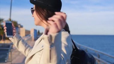 Siyah şapka ve güneş gözlüğü takan genç çekici kız blogger video aramasý konuşmak ya da bir cep telefonu ile ön kamera bir video El deniz kenarında kaydetme gülümsüyor. Ağır çekim. Denia, İspanya.