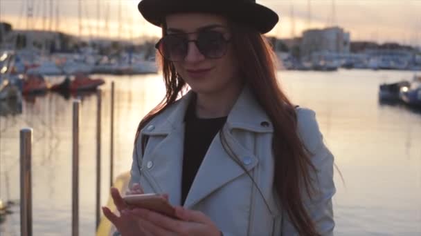 日落时分 年轻的微笑迷人的女孩博主戴着黑色帽子 戴着圆角太阳镜 在海上港口发短信或新的博客文章 手里拿着手机 慢动作 西班牙德尼亚 — 图库视频影像