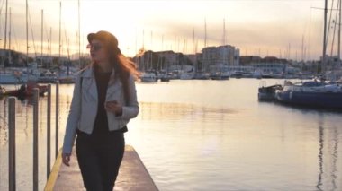 Mutlu genç çekici şık kız blogger bağlantı noktası deniz yürüyüş ve arka plan üzerinde teknelerle batımını cep telefonunu. Ağır çekim. Denia, İspanya.