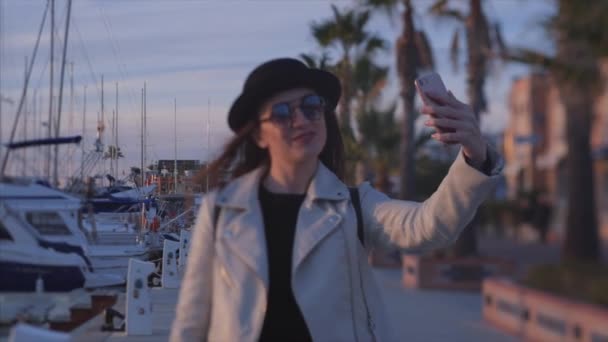 年轻迷人的女孩博主戴着黑色帽子 戴着太阳镜 在视频通话中微笑着说话 或者在前置摄像头上用手机在海边手里录制视频 慢动作 西班牙德尼亚 — 图库视频影像
