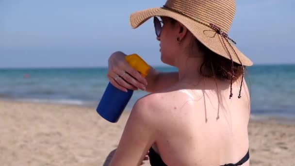 若い女性がビキニと麦わら帽子を身に着けているビーチで肩に日焼け止めを適用します 女の子青色の瓶から日焼け止めクリームを粉砕します ビデオ — ストック動画