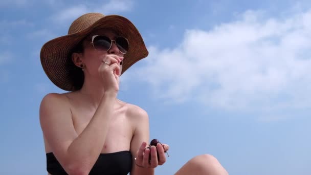 ビキニと青い空を背景に海のビーチでチェリーを食べて麦わら帽子を身に着けている若い女性 夏の暑い日 休暇の概念 — ストック動画