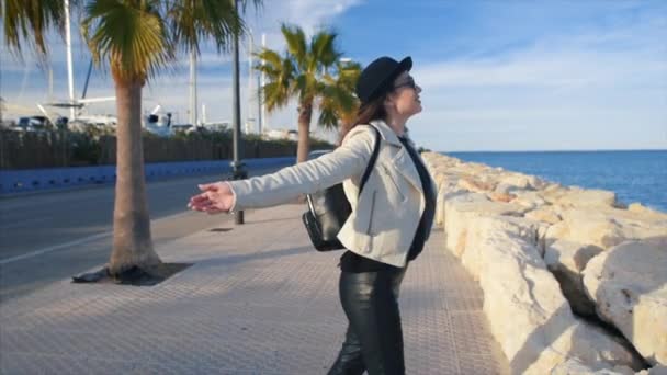 黒い帽子と丸いサングラスを身に着けている幸せな若い魅力的な女の子ブロガーは笑みを浮かべて 手に携帯電話を海に良い天気を楽しんでします デニア スペイン — ストック動画