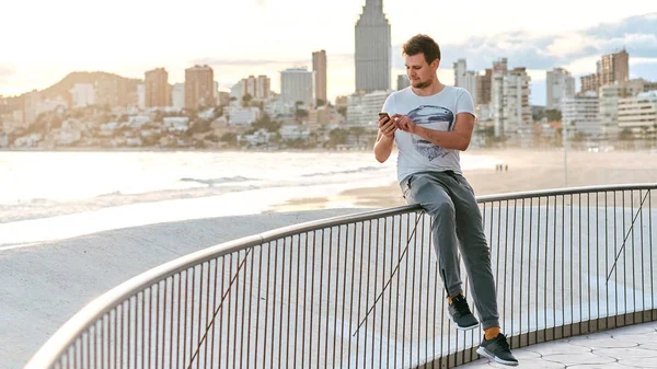 Jonge knappe man met smartphone tegen zee en stad panoramisch uitzicht — Stockfoto