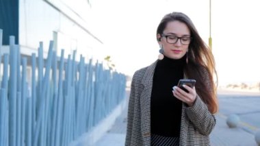 Gözlüklü ve elinde cep telefonu ile genç başarılı iş kadını yavaş hareket. Güneşli bir günde yürüyen şık başarılı kadın.