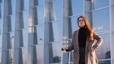 Kendine güvenen genç iş kadını cebindeki cep telefonunu çıkarıp havaalanından birini arıyor. Şık bir görünüm, zarif ceket, gözlük ve suicase. Taksiyi bekleyen iş kadını. 4k video.