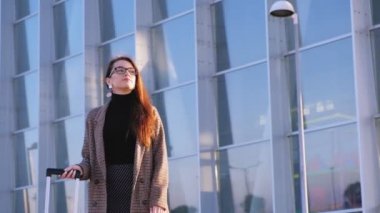 Kendine güvenen genç iş kadını havaalanında taksiye biniyor. Şık bir görünüm, zarif ceket, gözlük ve suicase. Taksiye koşan iş kadını. 4k video.