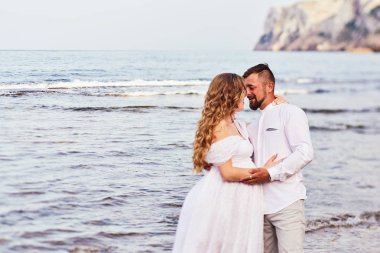 Genç hamile bir kadın kocasıyla kumsalda poz veriyor.