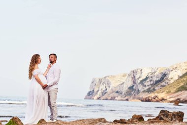 Genç hamile bir kadın kocasıyla kumsalda poz veriyor.