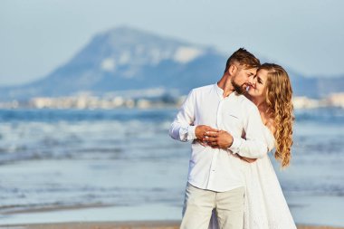 Beyaz gelinlikli genç sarışın gelin sahilde damadı kucaklıyor.