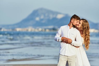 Beyaz gelinlikli genç sarışın gelin sahilde damadı kucaklıyor.