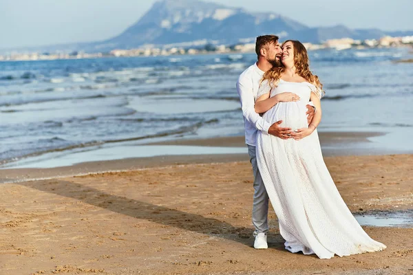 Młody mężczyzna przytula swoją ciężarną żonę w białej sukience na plaży — Zdjęcie stockowe
