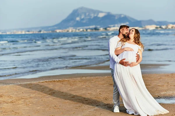 Молодий чоловік обіймає свою вагітну дружину в білій сукні на пляжі — стокове фото