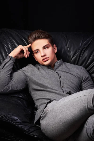 Fashion portrait of elegant young man sitting on leather sofa on black background