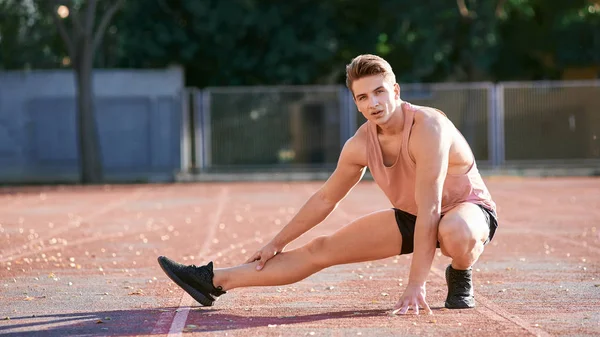 Young man runner stretching his leg on race track in stadium — Stockfoto