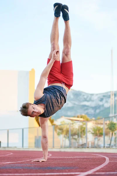 Young muscular man staying head over heels on one hand on the stadium