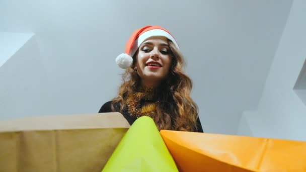 穿着黑色连衣裙和红色圣诞老人帽子的年轻女子 看着她五颜六色的购物袋 微笑着孤立在白色背景 黑色星期五 圣诞节或新年时间 慢动作 — 图库视频影像