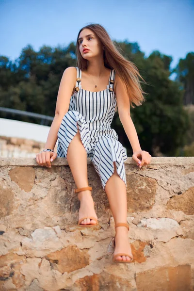 Mulher bonita vestindo macacão listrado e sandálias elegantes sentado em uma parede de pedra  . — Fotografia de Stock