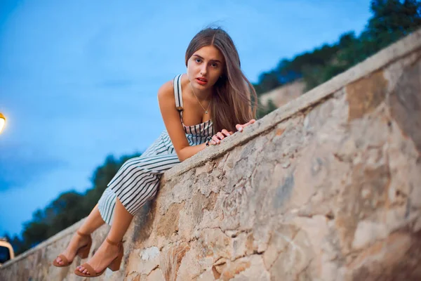 Belle femme portant des salopettes rayées et des sandales élégantes assises sur un mur de pierre  . — Photo