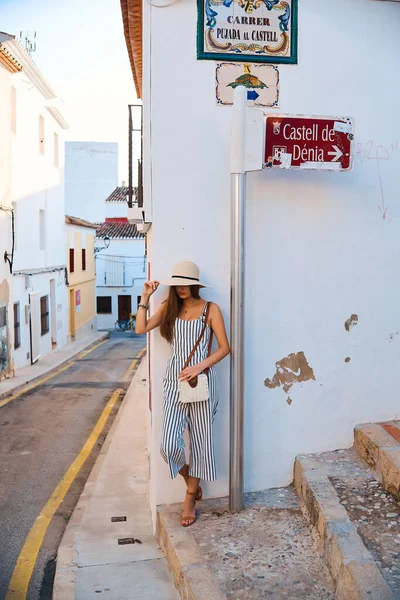 Mujer joven y elegante en sombrero de paja posando en la calle del casco antiguo contra la pared blanca . — Foto de Stock