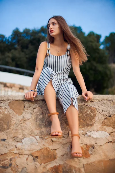 Mulher bonita vestindo macacão listrado e sandálias elegantes sentado em uma parede de pedra  . — Fotografia de Stock