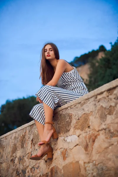 Mulher bonita vestindo macacão listrado e sandálias elegantes sentado em uma parede de pedra  . — Fotografia de Stock