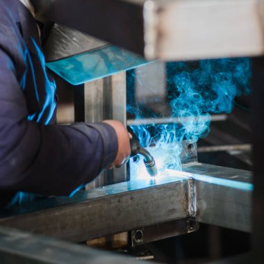 Yakın plan. Fabrikada mavi ceketli bir erkek işçi elinde kaynakla metal parçaları kaynak yapıyor. Kaynak yaptığı yerden beyaz, sönük bir duman çıkıyor.