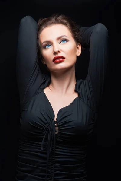 beautiful woman with beautiful brown hair in a dark dress looks up posing on a dark background, holding hands behind her head