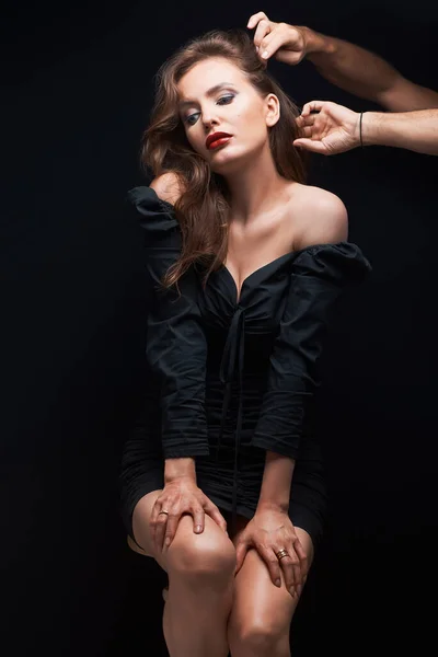 beautiful woman with beautiful brown hair in a dark dress on a dark background and male hands makes a hairstyle