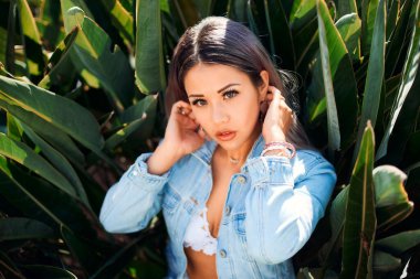 Sarışının güzel bir portresi, kot ceket ve açık mayo giymiş büyük yeşil yaprakların arka planında..